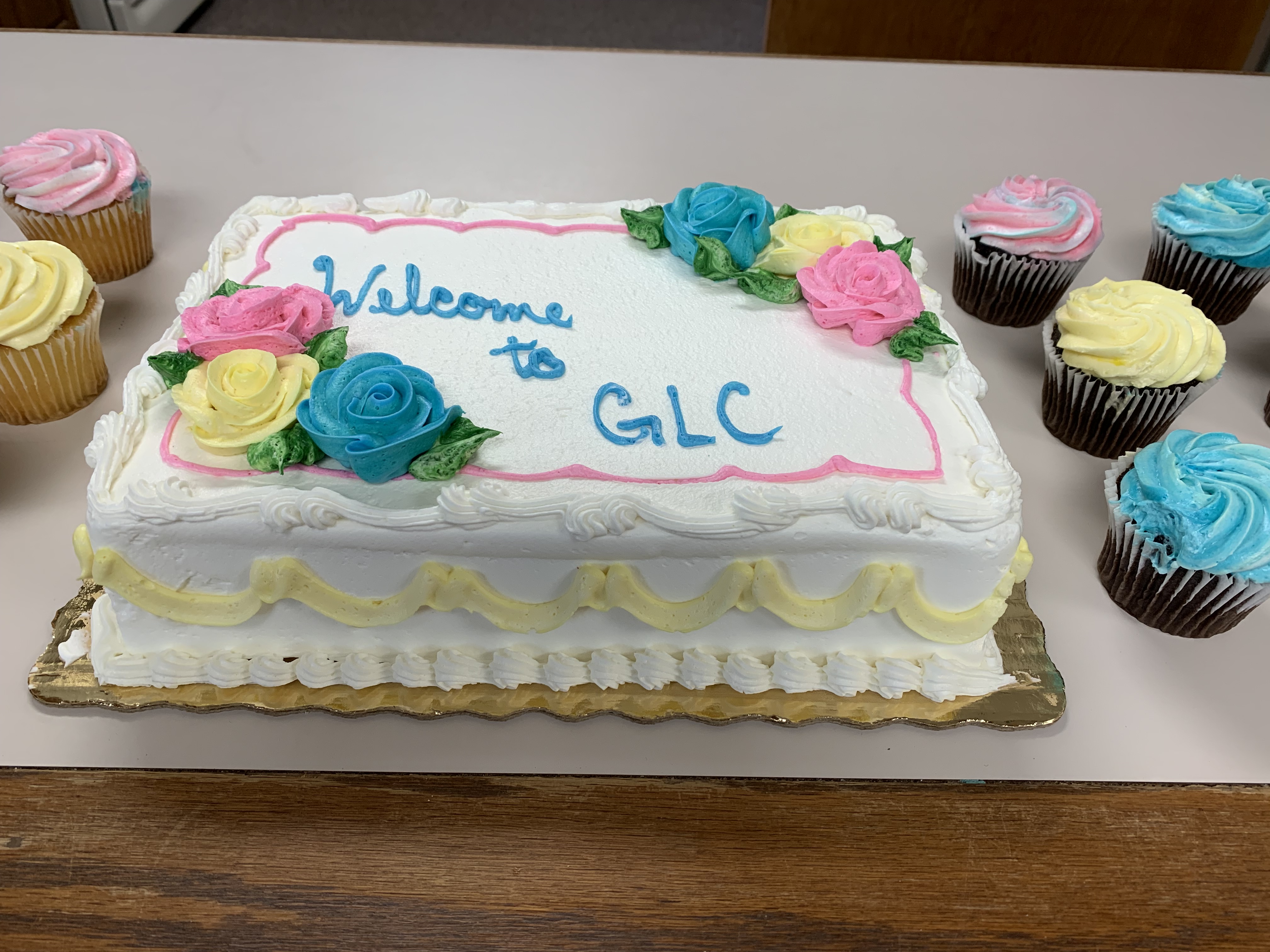 Pictured is a white frosted cake with blue, yellow and pink frosting flowers and the words "Welcome to GLC" written in blue frosting.