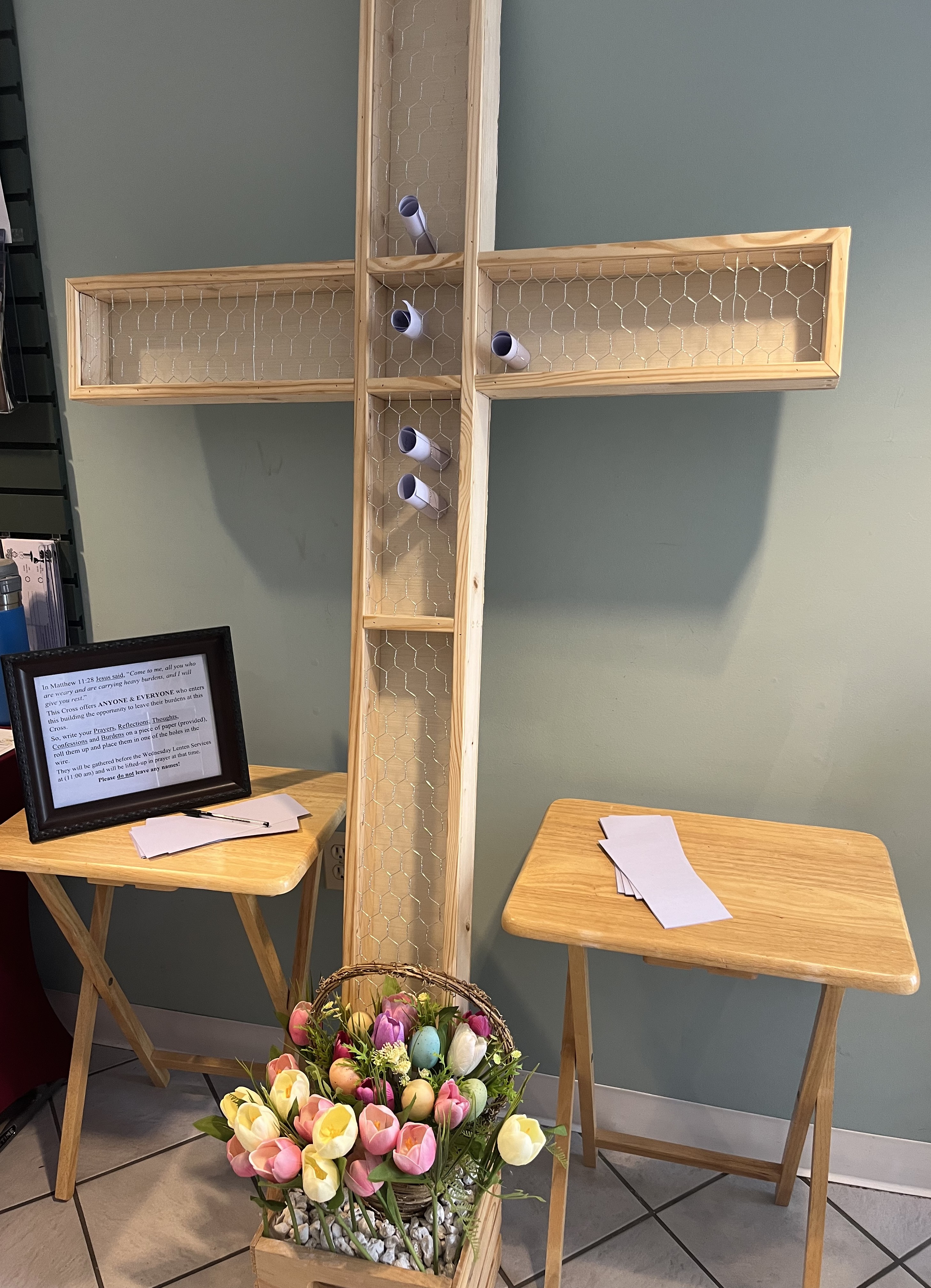 A wooden cross has a surface of chicken wire allowing for addition of prayer scrolls written on purple paper. There are blank scrolls available and a sign welcoming anyone to add prayers.